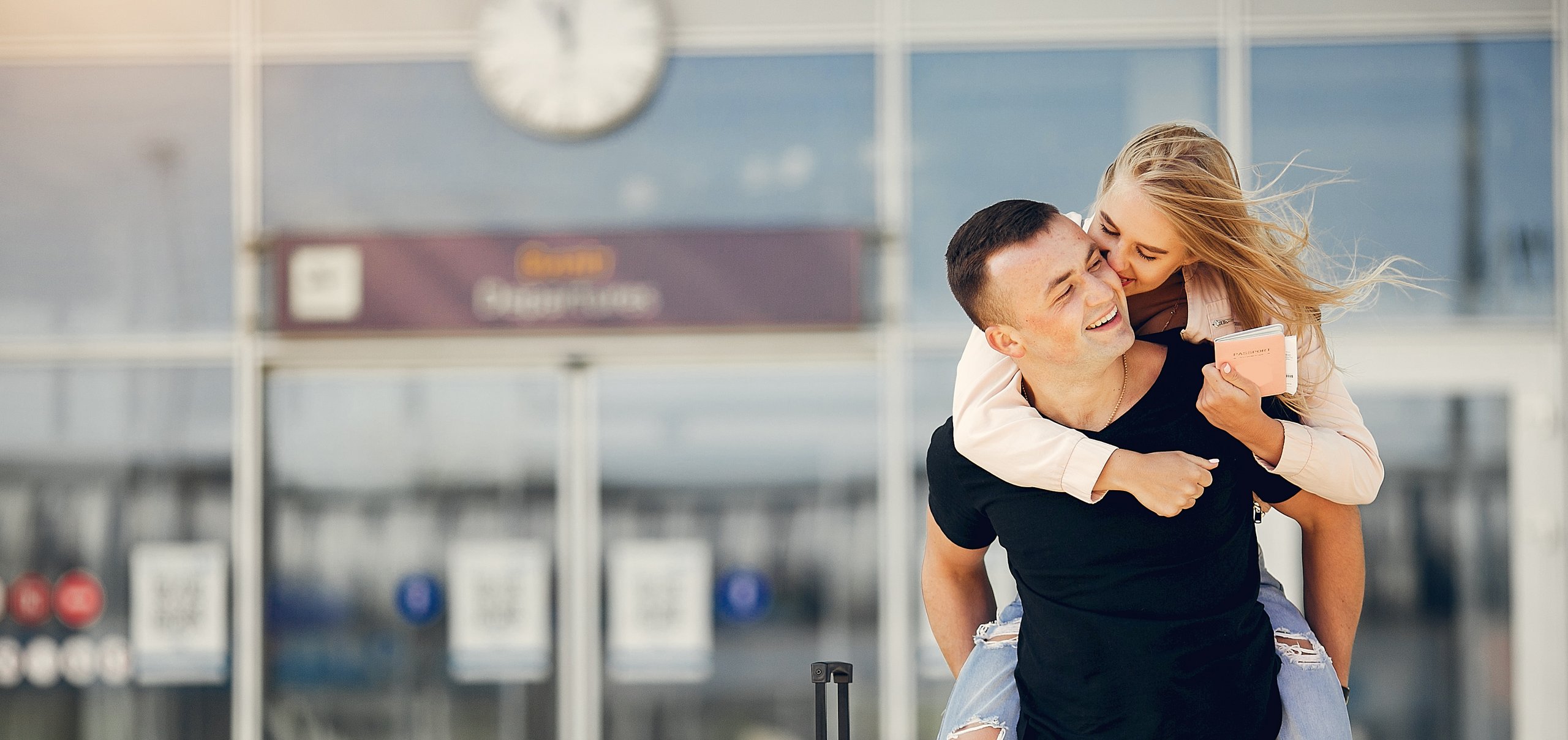 Mann trägt eine Frau auf dem Rücken. Sie hält ein Reisepass in der Hand. Beide lächeln