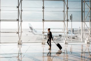 Man sieht ein Mann der einen Koffer hinter sich herzieht am Flughafen. Im Hintergrund ist ein Flugzeug zu sehen