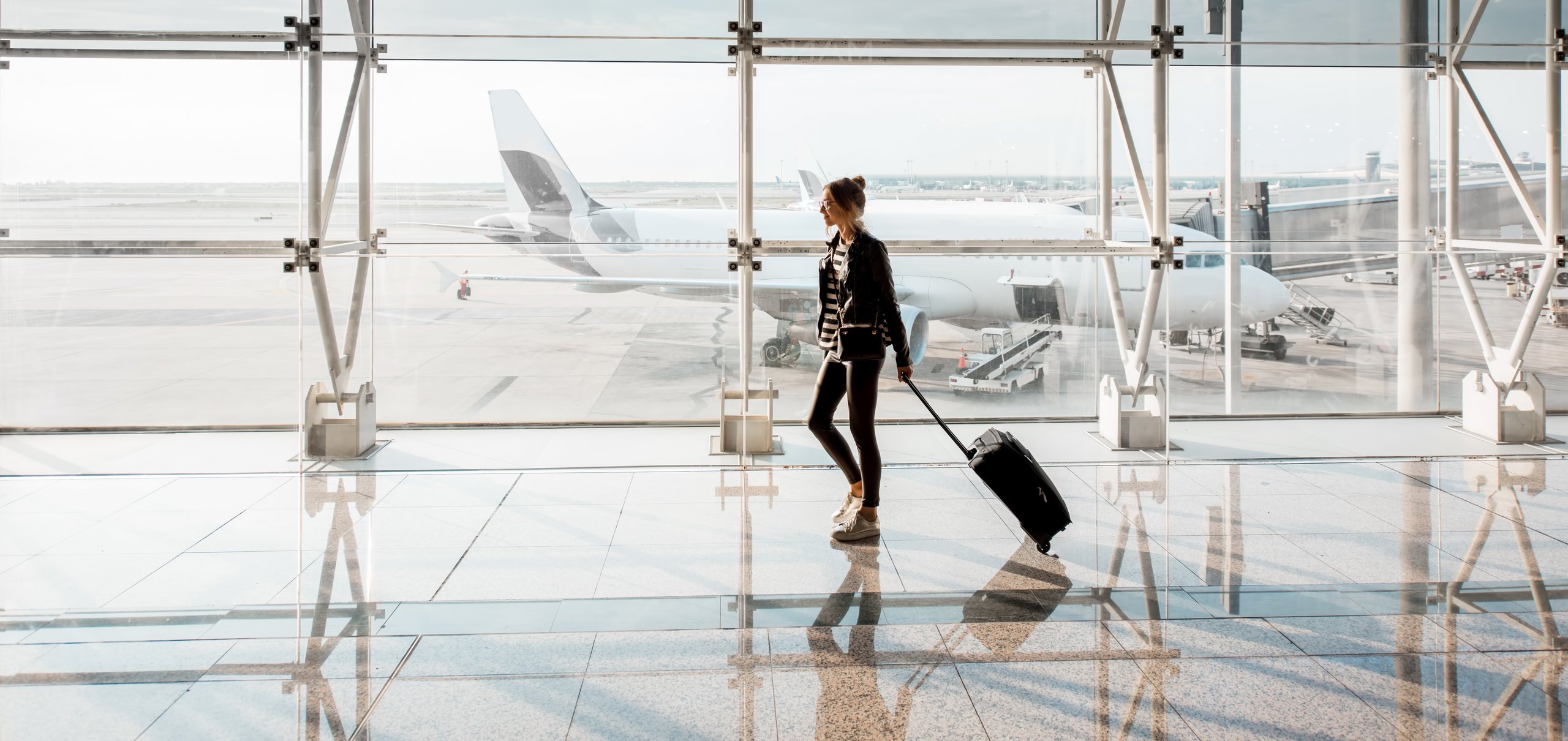 Man sieht ein Mann der einen Koffer hinter sich herzieht am Flughafen. Im Hintergrund ist ein Flugzeug zu sehen